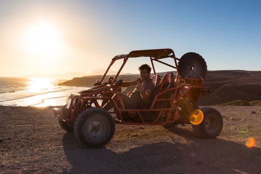 Active man driving quadbike on dirt road by the sea in sunset showing rocking sign to the camera.