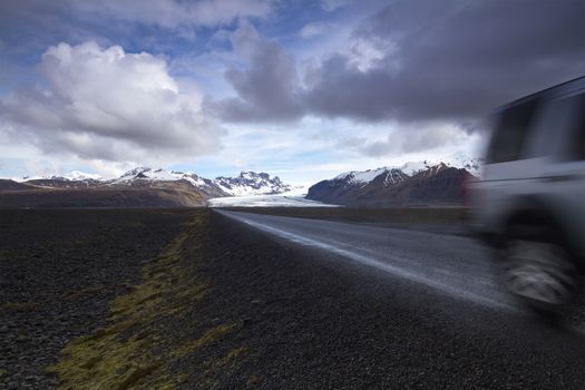 Horizontal photo of an off road car on a straight asphalt road coming from the mountains with clouds above and the Vatnajokull glacier in the background, Iceland