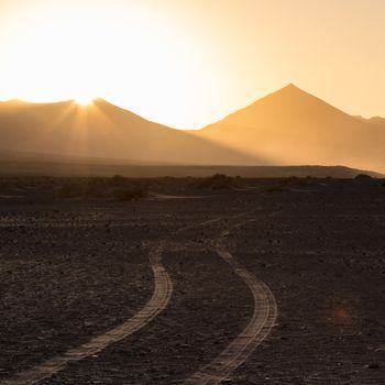 Car tire tracks in sand. Dirt road vanishing to the mountains sunlit in sunset. Copy space for text. Square composition.