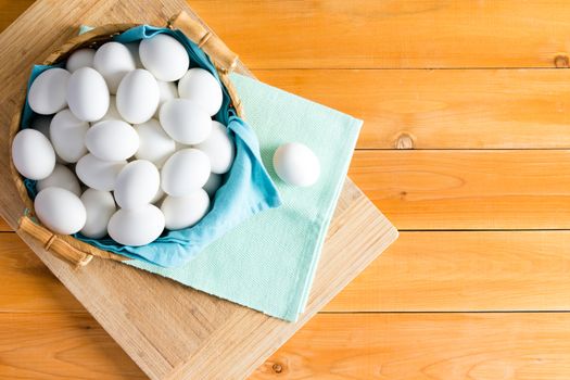 Basketful of fresh white eggs with one outside laid on a blue napkin, wooden background with copy space in an overhead view