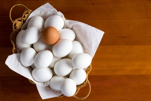 Wicker basket full of white eggs and single brown type on top over wooden table background with copy space on side