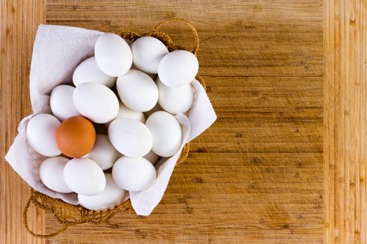 Basketful of clean white eggs displayed on a napkin with one brown one balanced on top in a conceptual image over a bamboo cutting board with copy space, overhead view