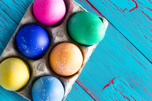 Six colored dyed Easter eggs in a cardboard egg box with copy space over a rustic exotic tropical blue wooden background with crackle paint, overhead view