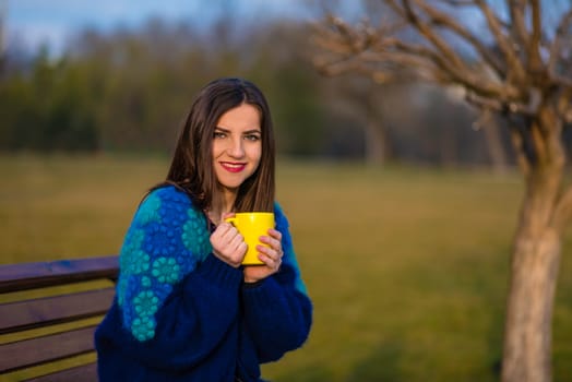 A teenager girl is sitting on a bench in a park and holding a yellow cup with two hands.