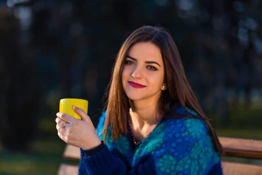 A teenager girl in turquoise blouse is sitting on a bench in a park and holding a yellow cup with two hands.