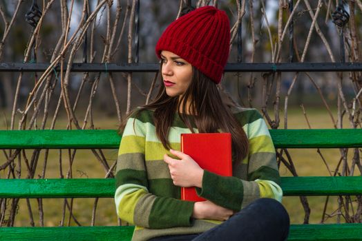 Teenager girl in red hat and green sweater sitting on a bench in a park and holding a red book.