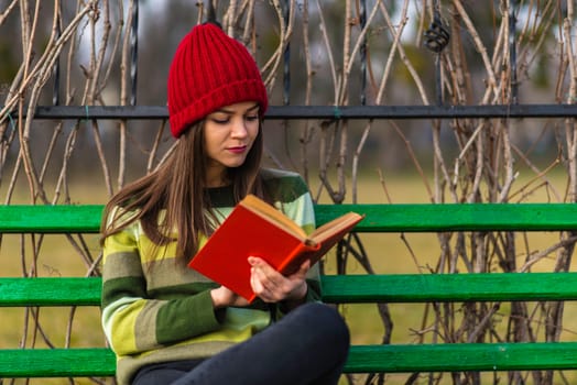 Teenager girl in red hat and green sweater sitting on a bench in a park and reading a book with red cover.