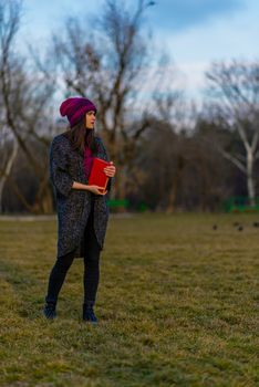 A teenager girl in purple hat holding book with red cover while standing on a lawn in a park and looking away.