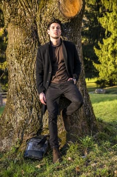 Handsome young man leaning against tree, looking at camera, in a sunny day wearing a black coat