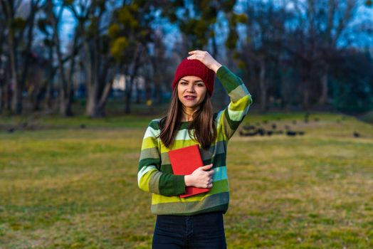 A teenager girl in red hat and green striped sweater holding book with red cover while standing on a lawn in a park