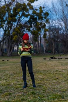 A teenager girl in red hat and green striped sweater and serious face is readin a book with red cover while standing on a lawn in a park.