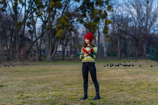 A teenager girl in red hat and green striped sweater and serious face is holding a book with red cover while standing on a lawn in a park and watches away.