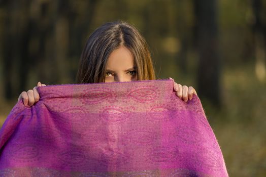 Behind purple scarf. Pretty teenager girl with brown eyes is covering her face with the purple scarf in the afternoon woods. Action takes place in autumn at afternoon in a forest.