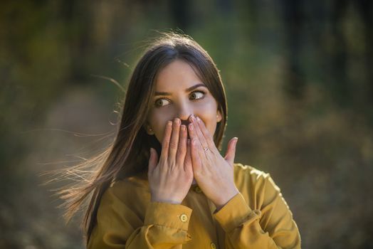 Amazed girl. Nice teenager girl having surprised expression on her face and covering her mouth with hands. Action takes place in an afterniin autumn forest. Girl is amazed by something from out of frame.