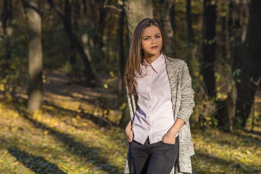 A pretty teenager girl is posing in an autumn oak forest at afternoon. She has a detached and thoughtful expression on her face. She is holding her hands in her jeans pockets. 