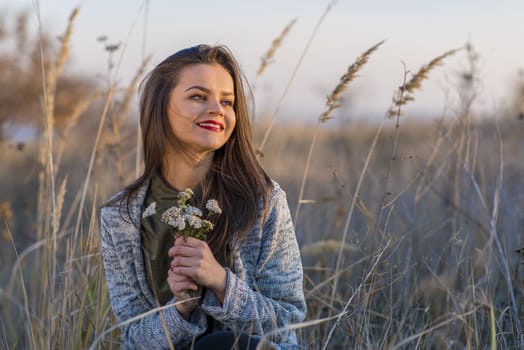 Looking at sun. A beautiful smiling teenage girl sitting in a field with a bouquet of field flowers in hands and looking towards autumn sunset. Girl has brown eyes and hair and red lips. 