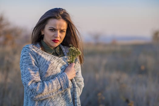 Getting cold in a field. A teenager girl wrapped in a warm jacket in a field during sunset. She is holding a bouquet of field flowers in her hand. She has a sad expression on her face. Girl has red lips and half of her face is covered with light and the other one is in the shadow.