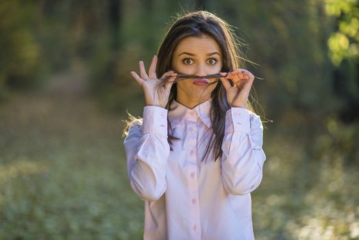 A teenage girl is making mustache from her hair with eyes wide open and a quirky expression on her face.