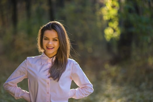 Happpy teenage girl is posing in a forest at afternoon in autumn. She is wearing a pink shirt and her hair is backlit by the sun. She has a big happy smile on her face and pleased expression.