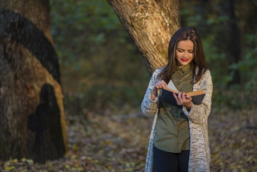 Fairy tales in afternoon. Smiling by reading fairy tales. A teenager girl is reading a book in an evening forest with two oak trees in the background. She casts shadow on one of the trees. The girl has a happy expression on her face.
