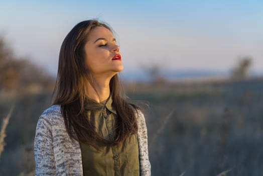 Enjoying sun before winter. A beautiful teenage girl standing in a field at autumn sunset, enjoying last sun days before winter. Girl has brown hair and red lips.