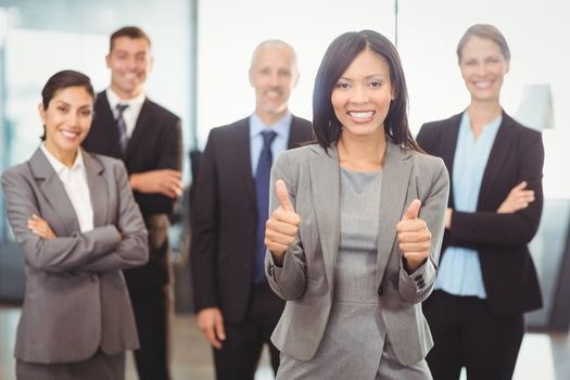 Portrait of businesswoman giving thumbs up and colleagues standing behind with arms crossed in office