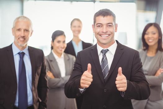 Portrait of businessman giving thumbs up and colleagues standing behind with arms crossed in office