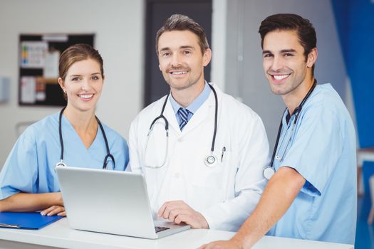 Portrait of smiling doctor and colleagues with laptop while standing at desk in hospital