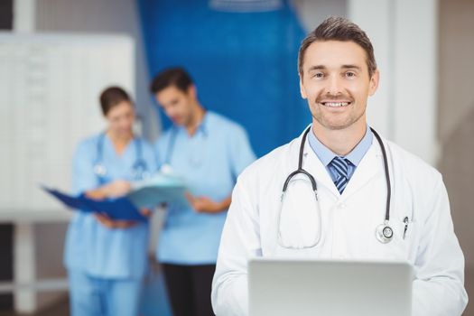 Portrait of smiling doctor with laptop and colleagues standing on background