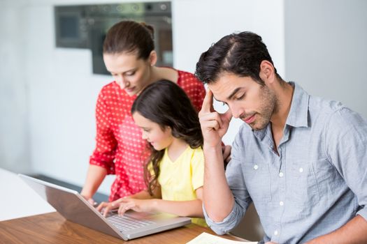 Daughter and mother working on laptop with tensed father sitting at desk in home