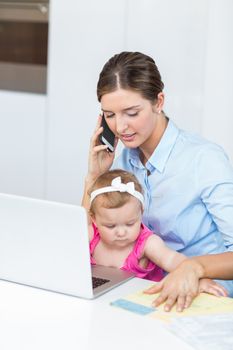 Woman talking on mobile phone while sitting with baby girl by laptop at home