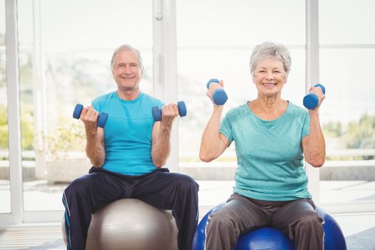 Portrait of smiling senior couple holding dumbbells while sitting on exercise ball at home