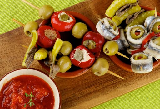 Delicious Spanish Snacks with Stuffed Small Peppers, Anchovies, Tomatoes Sauce, Green Olives and Bread Sticks in Various Bowls closeup on Wooden Cutting Board. Top View