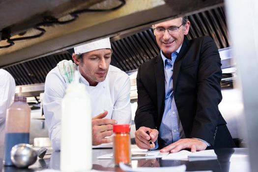 Male restaurant manager writing on clipboard while interacting to head chef in commercial kitchen