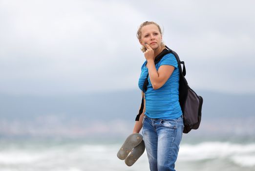 Pretty young blonde woman in a blue t-shirt and jeans with a backpack standing on a sea background.  Shallow depth of field. Focus on model.