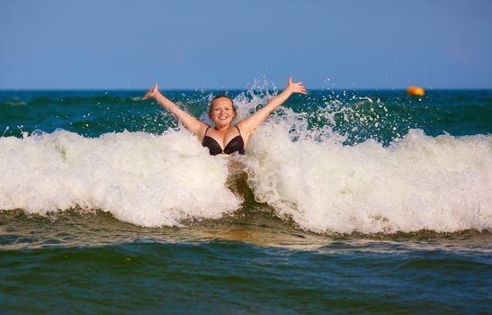 Beautiful smiling girl with her hands raised in the foam of a sea wave. Happy young woman. Bright sunny day.