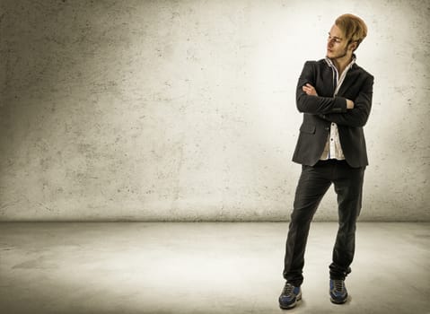 Handsome blond young man standing against concrete wall, looking away to a side, wearing white shirt and jacket. Full length shot