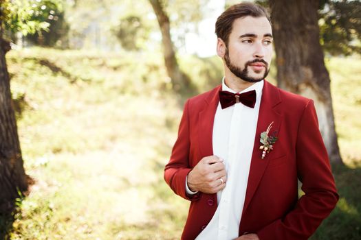 Stylish groom in tuxedo looking away suit marsala red, burgundy bow tie. Man stick to the edge of his jacket, outdoors. Professional hairstyle, beard, mustache. Wedding preparations, getting ready. Copy space for text. Celebration.