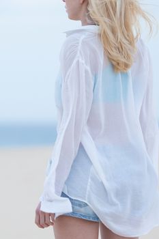 Sensual blonde woman wearing white loose casual shirt on vacations looking down sandy beach in dusk.