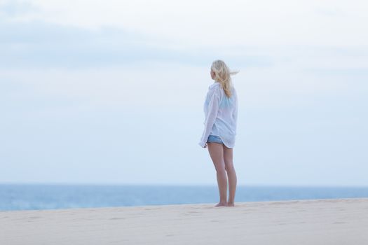 Meditative, sensual blonde woman wearing white loose casual shirt on vacations looking at horizon at dusk.