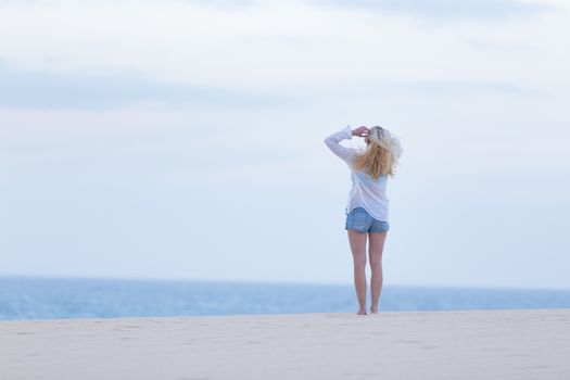 Meditative, sensual blonde woman wearing white loose casual shirt on vacations looking at horizon at dusk.