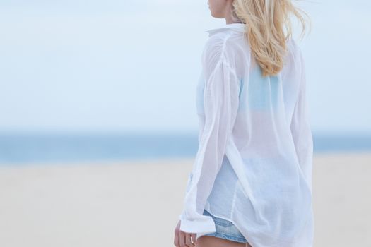 Sensual blonde woman wearing white loose casual shirt on vacations looking down sandy beach in dusk.
