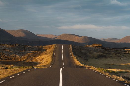 Horizontal photo at sunset of a straight asphalt road going into the mountains with clouds above in Reykjanes peninsula, Iceland