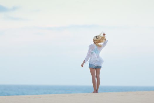 Meditative, sensual blonde woman wearing white loose casual shirt on vacations, looking at horizon, enjoying peaceful early morning.