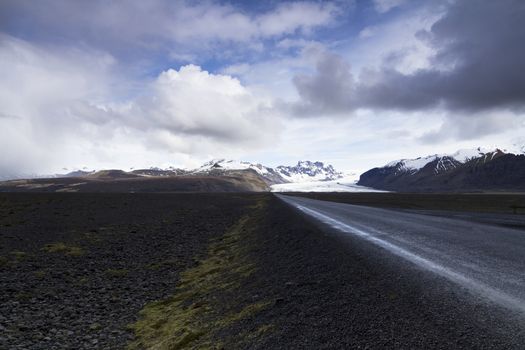 Horizontal photo of a straight asphalt road coming from the mountains with clouds above and the Vatnajokull glacier in the background, Iceland