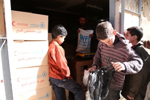 SYRIA, Saqba: A child carries a food pack as Syrian Red Crescent staff survey the delivery of humanitarian aid at a warehouse in Saqba, in the rebel-held Eastern Ghouta area, southern Syria, on the outskirts of the capital Damascus on March 15, 2016 during an operation in cooperation with the UN to deliver aid to thousands of besieged Syrians. UN aid convoys head to two Syrian towns besieged by government forces to deliver life-saving food and medicine to some 30,000 people, a UN spokesman said. 