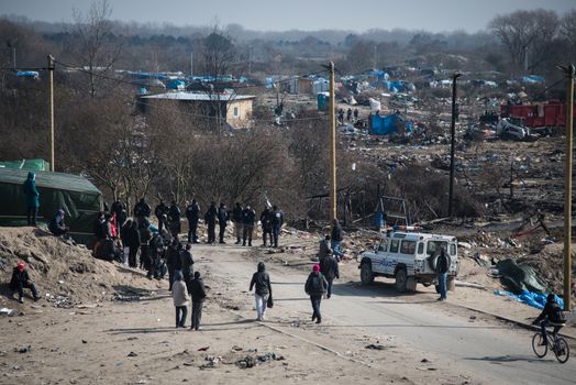 FRANCE, Calais: Migrants walk past French anti-riot policemen standing guard during the dismantling of the southern part of the so-called 'Jungle' migrant camp in Calais, northern France, on March 14, 2016.Thousands of migrants have been living in the Jungle and other smaller camps along the northern coast, desperate to reach Britain where many have family or community ties and see better hopes of gaining employment or education. 