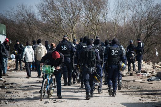 FRANCE, Calais: Migrants walk past French anti-riot policemen standing guard during the dismantling of the southern part of the so-called 'Jungle' migrant camp in Calais, northern France, on March 14, 2016.Thousands of migrants have been living in the Jungle and other smaller camps along the northern coast, desperate to reach Britain where many have family or community ties and see better hopes of gaining employment or education. 