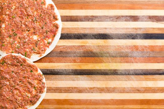 Two prepared Turkish lahmacun with a savory minced meat and veggie mix on a dough base ready to cook forming a side border on a decorative striped cutting board with copy space, close view from above