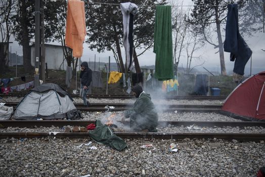 GREECE, Idomeni: A man warms along the railway tracks next to tents at the Idomeni refugee camp on the Greek Macedonia border on March 15, 2016 in Idomeni, Greece. Yesterday, an attempt by some of the residents of the camp to cross the border to Macedonia was thwarted by Macedonian security forces who returned the migrants to Greece. The decision by Macedonia to close its border to migrants last week has left thousands of people stranded at the Greek transit camp. The closure, following the lead taken by neighbouring countries, has effectively sealed the so-called western Balkan route, the main migration route which has been used by hundreds of thousands of migrants to reach countries in western Europe, such as Germany. Humanitarian workers have described the conditions at the camp as desperate, which has been made much worse by recent spells of heavy rain.